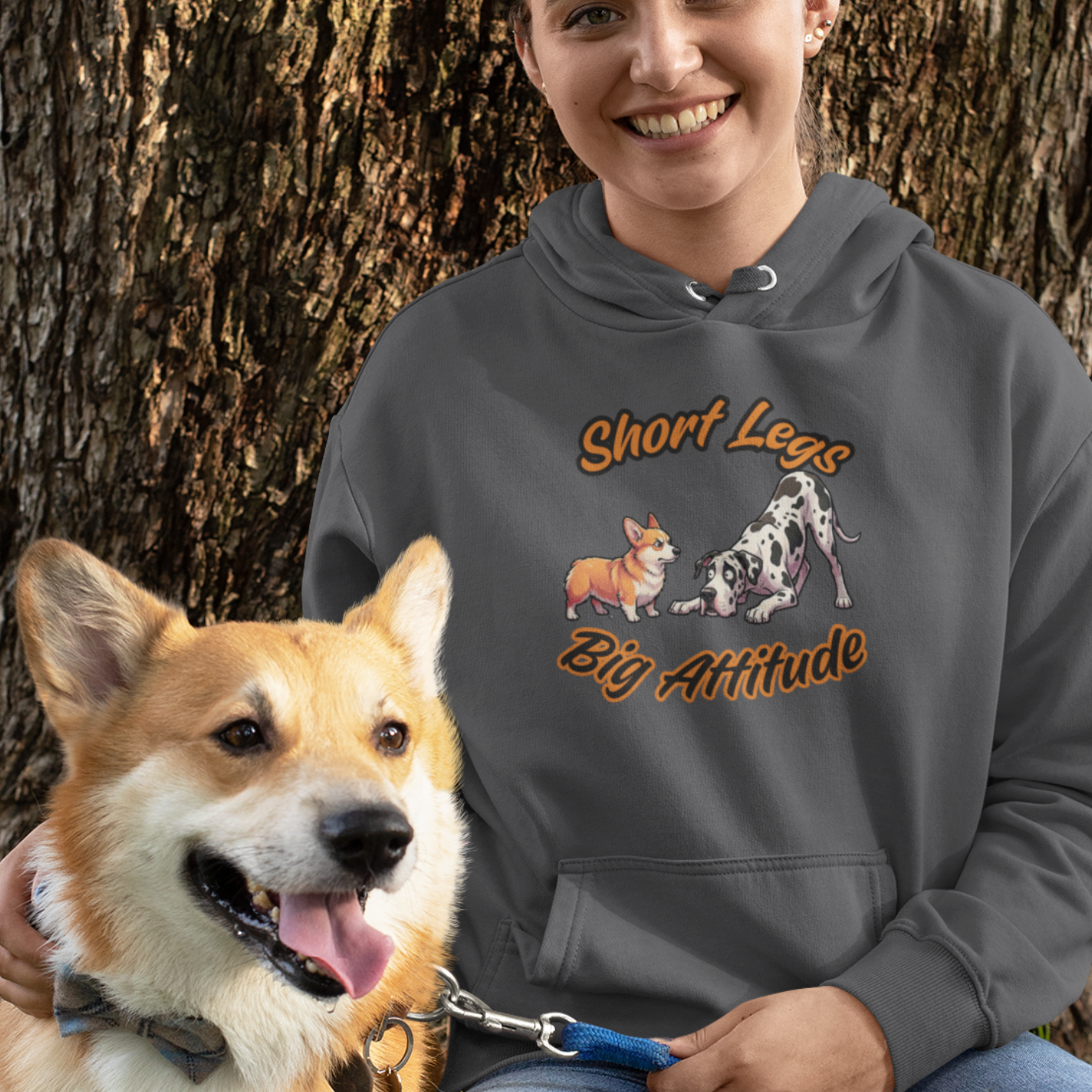 Woman wearing a hoodie with dog graphics and text, sitting next to a corgi in a park.
